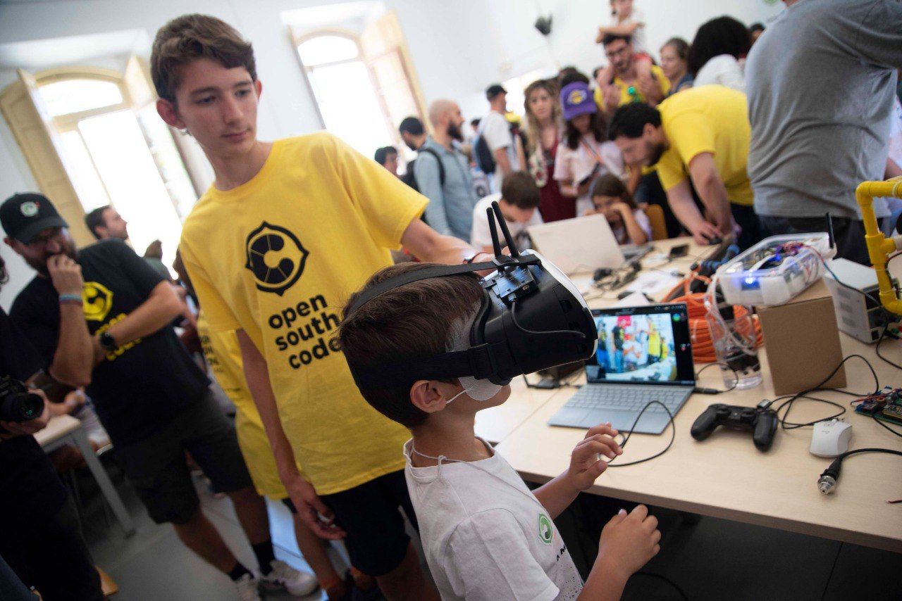 Vista de los talleres prácticos de OpenSouthKids, con un niño probando una experiencia en realidad virtual mientras un adolescente colaborador en el evento le muestra cómo funciona la tecnología.